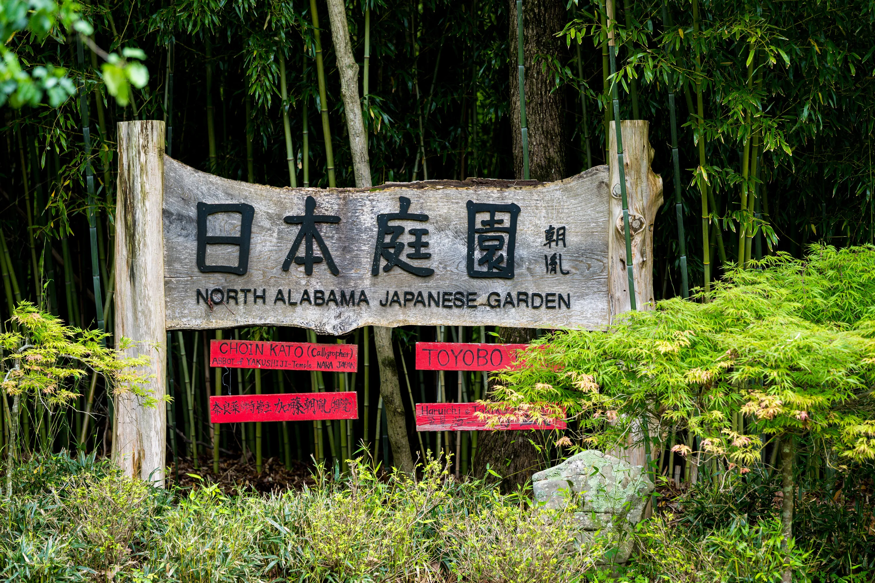 Weathered wooden entrance sign reading 日本庭園 (Nihon Teien) — North Alabama Japanese Garden, framed by bamboo and Japanese maple